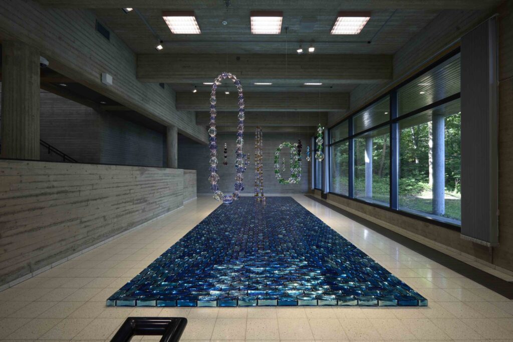 Sininen joki, 2024. intialainen peililasi, 12 x 1100 x 352 cm. Yläpuolella teoksia Helminauha- ja Rakastaja-sarjoista, 2024, muranon lasia ja ruostumatonta terästä.